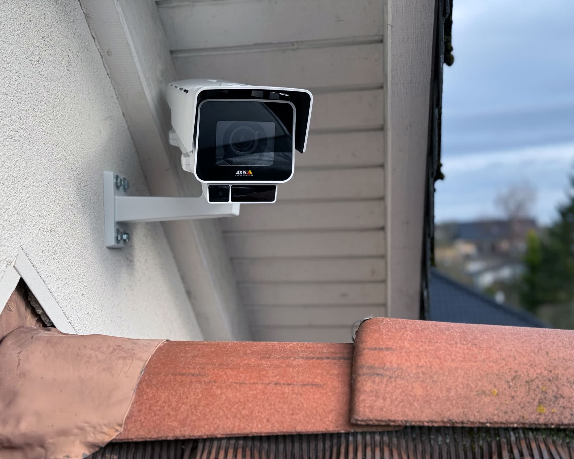 Weiße AXIS-Überwachungskamera unter einem Dachüberstand an einem Wohnhaus montiert.